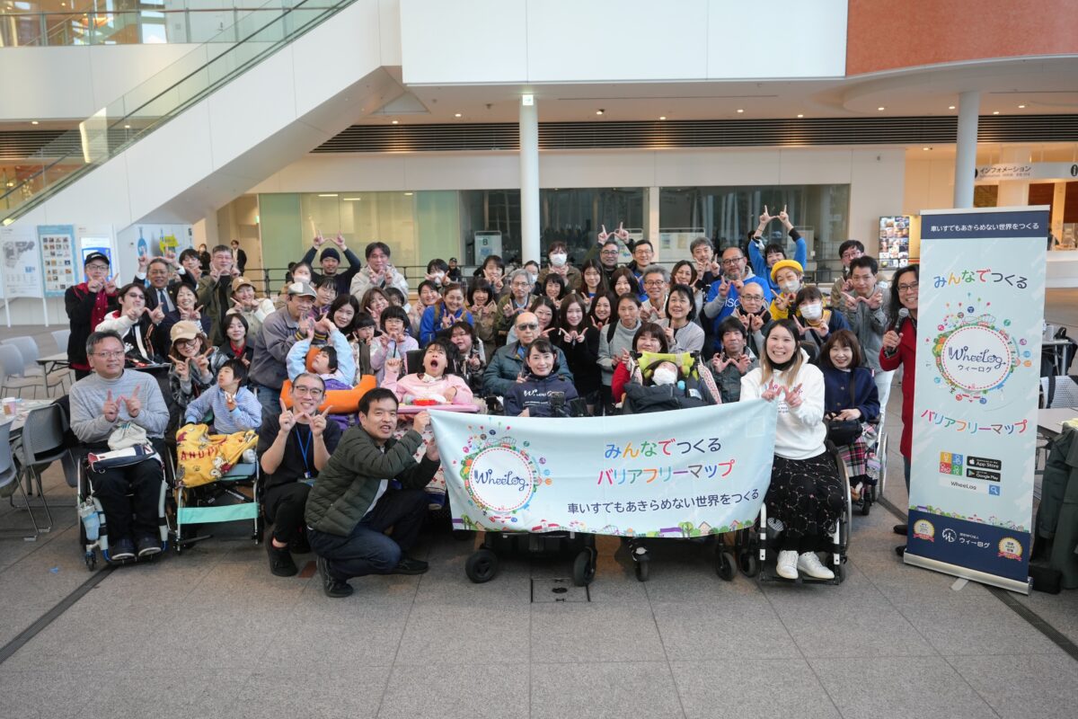 横浜市役所アトリウムでの集合写真。「みんなでつくるバリアフリーマップ」と書かれた横断幕を囲み、車いすユーザーを含む総勢60名の参加者が、笑顔で指差しポーズ（WheeLog!ポーズ）をとっている。
