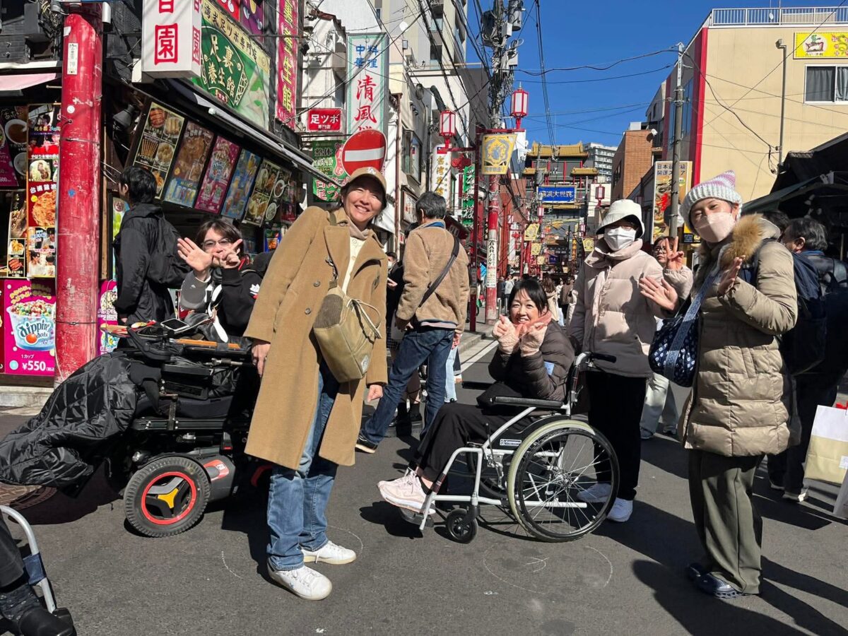 晴天の横浜中華街。カラフルな看板が並ぶ通りで、車いすユーザーとサポートする仲間たちが、リラックスした笑顔で写真に収まっているスナップショット。